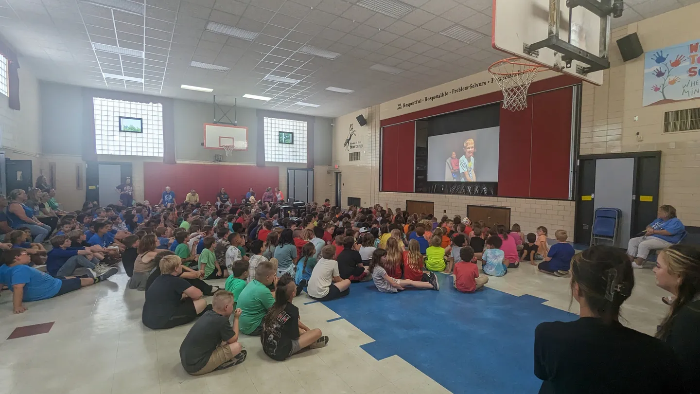 Students and adults gathered in a school gym to watch a presentation.